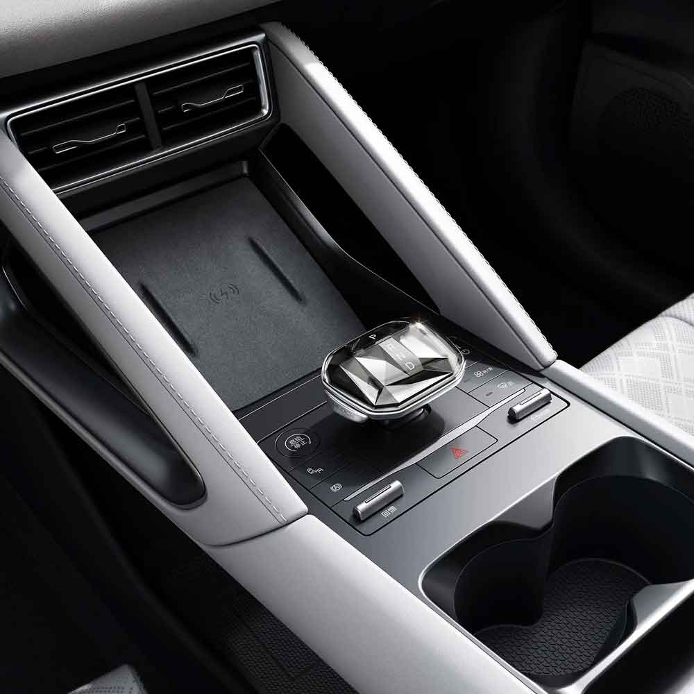 Close - up of a car's center console, featuring a crystal - like gear shifter, wireless charging pad, and cup holders.