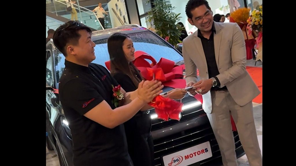 People at a car dealership ceremony, with one person in a suit handing over something to another in front of a car adorned with a big red bow.
