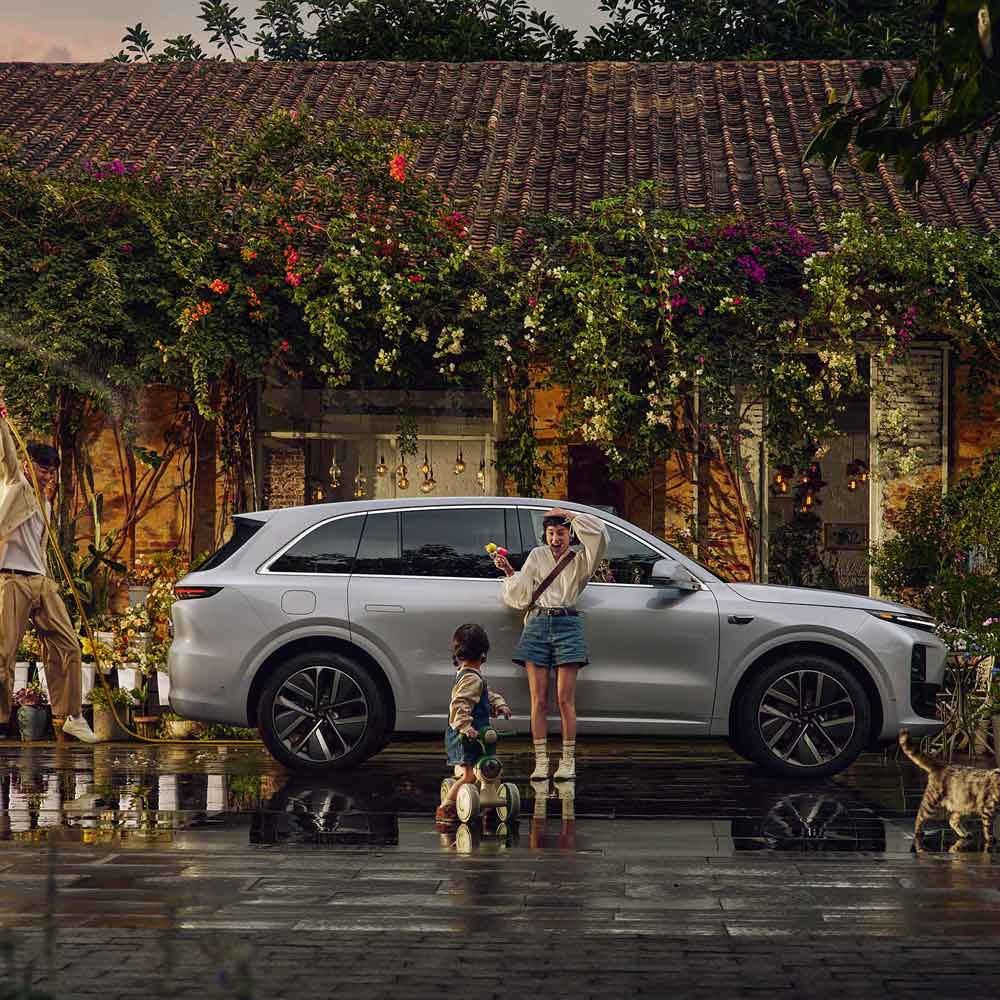A silver SUV parked in front of a quaint, vine - covered house, with a woman, a child, and a man (watering plants) in the scene.