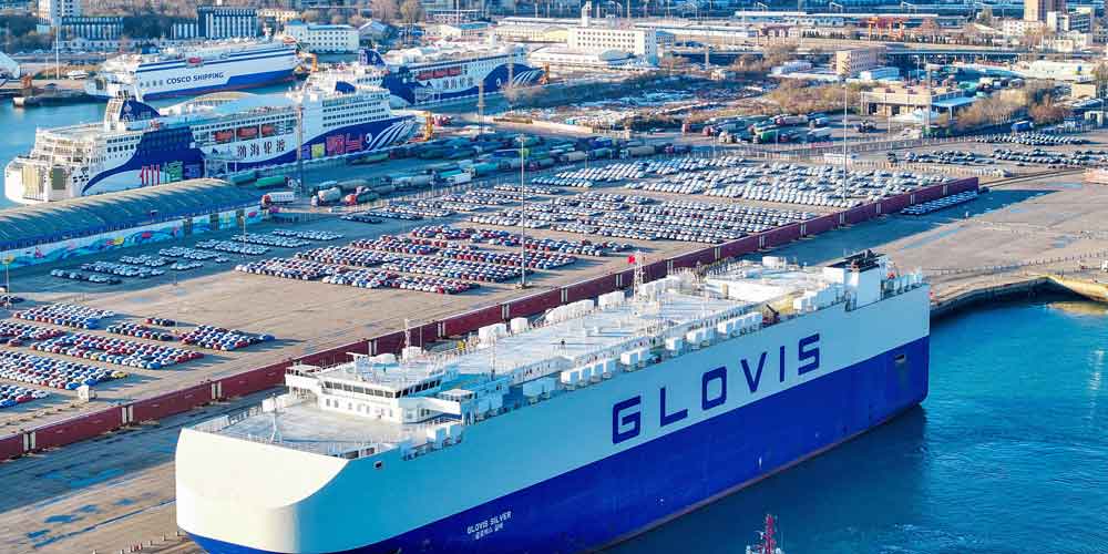 ទិដ្ឋភាពពីលើអាកាសនៃកំពង់ផែមួយ ដែលមានកប៉ាល់ដឹកជញ្ជូនរថយន្តធំដែលមានឈ្មោះ "GLOVIS" និងរថយន្តជាច្រើន។