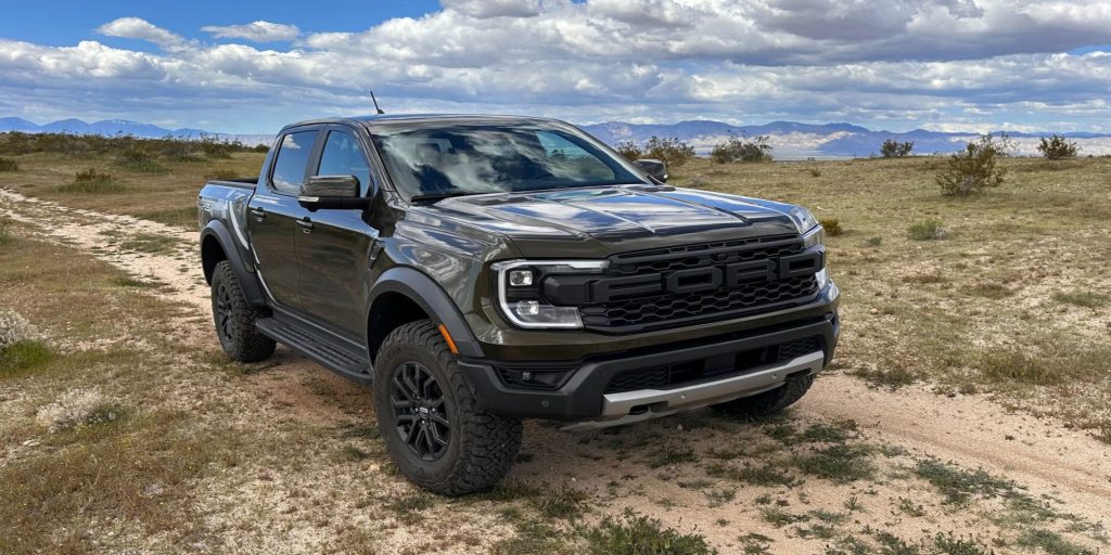 A Ford truck parked on a grassy and gravelly field under a blue sky with clouds, with mountains in the distance.