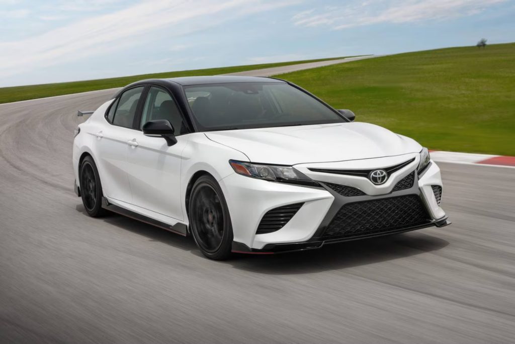 A white Toyota sedan driving on a racetrack with a green field and blue sky in the background.