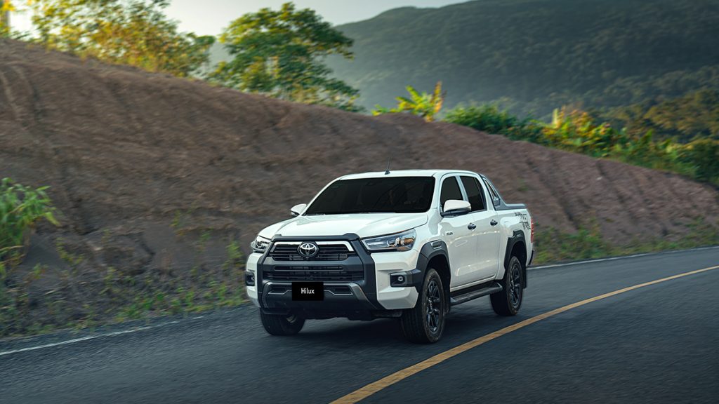 A white Toyota Hilux pickup truck driving on a road with a hilly, vegetated landscape in the background.
