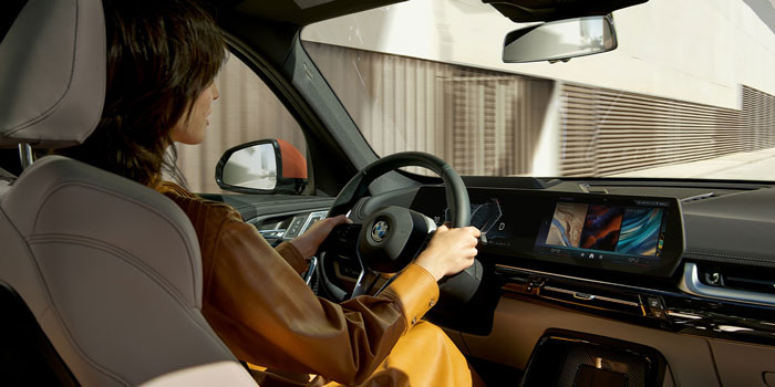 2025 BMW X1: tan leather interior, large touchscreen, navigating with steering wheel—city drive scene.