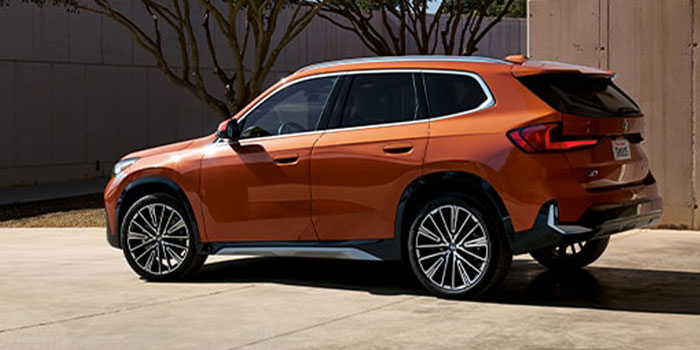 Orange 2025 BMW X1: sleek side design, alloy wheels, parked by concrete wall and trees.