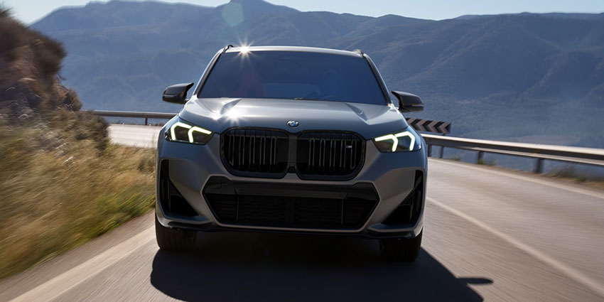 2025 BMW X1: front view, driving on mountain road, illuminated headlights, scenic backdrop.