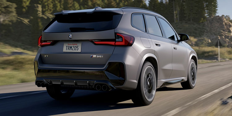 Gray BMW X1 M35i: rear view, driving on rural road, mountainous landscape in background.