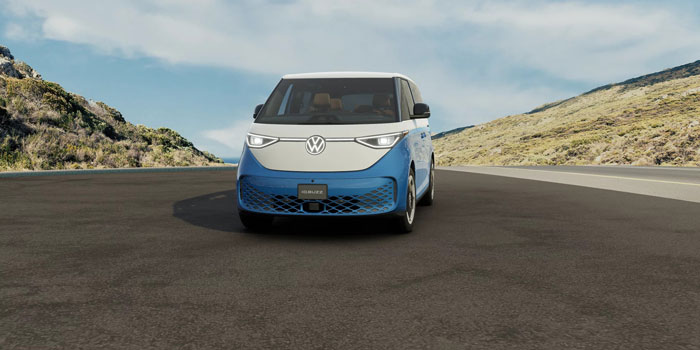 Front view of a 2025 Volkswagen ID. Buzz on a road, set against hilly, vegetated landscape under a partly cloudy sky.