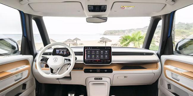 2025 Volkswagen ID. Buzz's dashboard: white/ beige interior, digital screens, steering wheel; beach view through front windows.