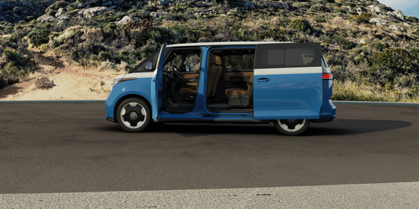 2025 Volkswagen ID. Buzz with open side doors, showing brown interior seats; rocky, vegetated hills in the background.