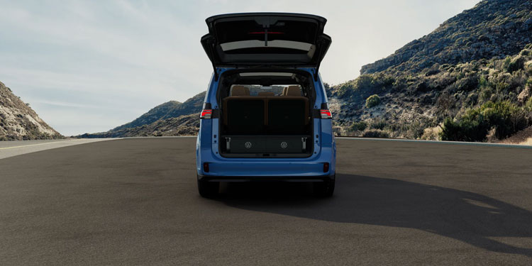 Rear view of a 2025 Volkswagen ID. Buzz with open trunk, showing interior storage; mountainous landscape behind on a paved road.