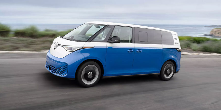 A blue-and-white Volkswagen ID. Buzz van drives along a coastal road, with the ocean and cliffs in the background.