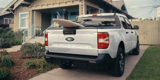 Rear view of a 2026 Ford Maverick pickup with a paddle board sticking out of the truck bed parked in front of a home