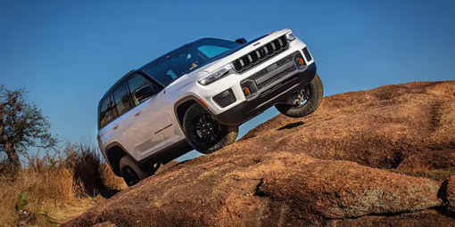 Display A white 2026 Jeep Grand Cherokee Trailhawk 4xe, crawling up a rock hill in the desert