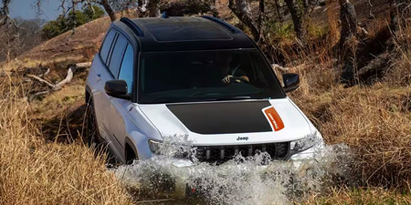 Display A white 2026 Jeep Grand Cherokee Trailhawk 4xe with black hood insert, entering a shallow stream from a grassy trail, off-road