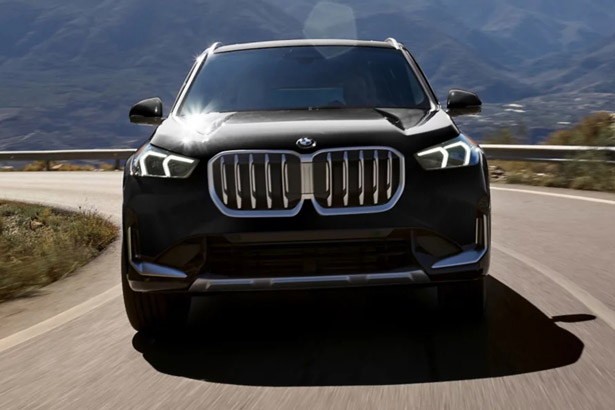 Front view of an 2026 BMW X1 driving on a curvy road on a sunny day with mountains in the background.